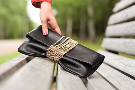 Woman hands is picking up a black leather clutch bag from a bench.