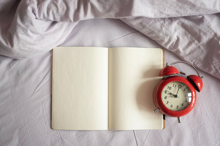 Blank notebook with empty pages for copy space and red alarm clock on the bed blanket background.の写真素材