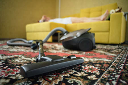 Overworked tired woman is lying on the sofa with a vacuum cleaner on the foreground.の写真素材