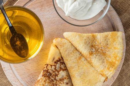 Thin lace Pancakes in stack homemade pastries on wooden board close-up with honey and sour cream. Food concept holiday Shrovetide Maslenitsa Top view flat lay copy space close-upの写真素材