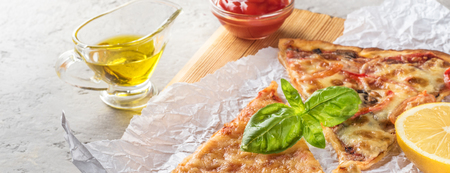 Two pieces of pizza with mushrooms and cheese and also with lemon slices and basil leaves on a wooden board covered with baking paper, served with tomato sauce and olive oil on a gray concrete stone backgroundの写真素材