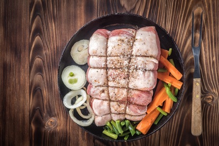 Raw pork chop marinated meat roll with coarse sea salt pepper assorted vegetables with carrots green onions and onion rings in cast iron skillet On wooden table background served with meat fork.の写真素材