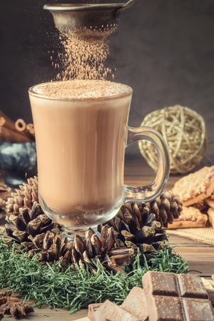 Glass cocoa mug or coffee with milk froth on wooden table served cookies cinnamon chocolate and winter holiday accessories. New Year Christmas card. Hand pours of cocoa powder.の写真素材
