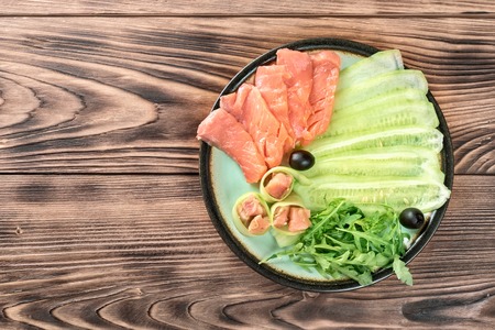 Plate with sliced pieces of red salmon fish, cucumber slices, arugula olives and rolls on wooden table. Concept of Mediterranean diet, nutrition, organic productsの写真素材