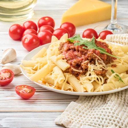 Italian pasta bolognese penne rigatone minced meat in tomato sauce and parmesan cheese. Still life on white wooden table served with cherry tomatoes, carafe of olive oil and glass of white wineの写真素材