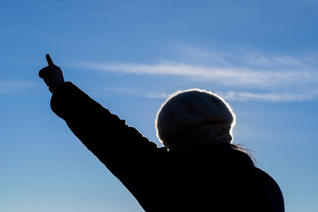 Silhouette of a young woman raised hand with her forefinger against sky background.の写真素材