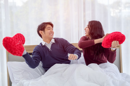 Smiling young asian couple on white bed with red heart-shaped pillow,  Valentine's day and happiness concept.の写真素材