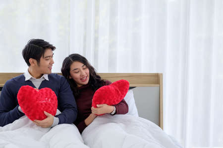 Smiling young asian couple on white bed with red heart-shaped pillow,  Valentine's day and happiness...