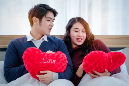 Smiling young asian couple on white bed with red heart-shaped pillow,  Valentine's day and happiness concept.の写真素材