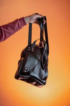 A woman's hand holds a leather backpack against an orange background