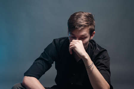 Sad young man in black shirt sits on a gray background with smoke blueの写真素材