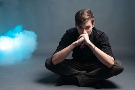 Sad young man in black shirt sits on a gray background with smoke blueの写真素材