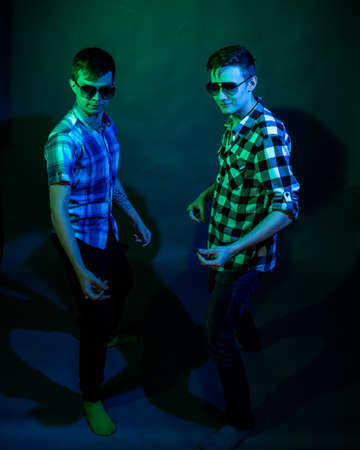 Two young men in plaid shirts dance in the studio with multicolored light
