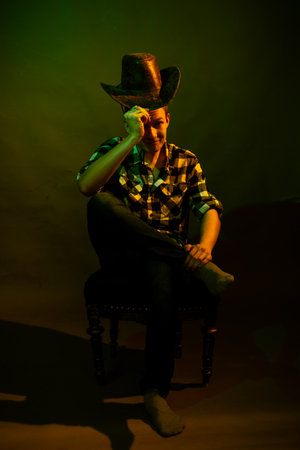 A young man in a plaid shirt with a cowboy hat sits on a chair, illuminated by a green and yellow lightの写真素材
