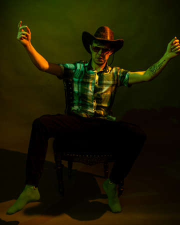 A young man in a plaid shirt with a cowboy hat sits on a chair, illuminated by a green and yellow lightの写真素材
