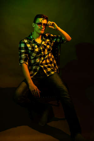 A guy in a plaid shirt and glasses sits on a chair illuminated by multicolored light against a dark background.の写真素材