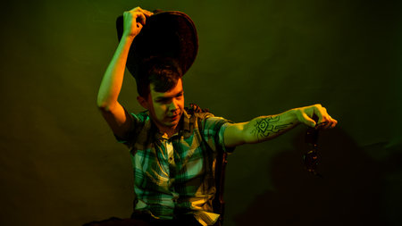 A young man in a plaid shirt with a cowboy hat sits on a chair, illuminated by a green and yellow lightの写真素材