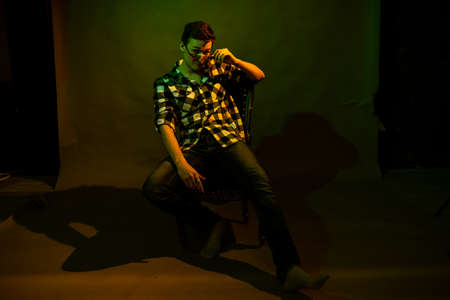 A guy in a plaid shirt and glasses sits on a chair illuminated by multicolored light against a dark...