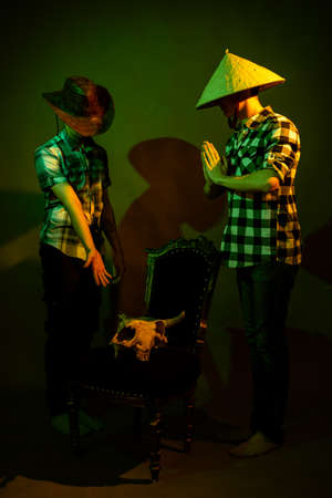 Two guys in plaid shirts and hats stand by a chair with a bull skull illuminated by multicolored lightの写真素材