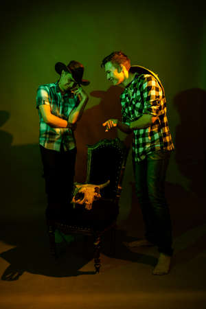 Two guys in plaid shirts and hats stand by a chair with a bull skull illuminated by multicolored lightの写真素材