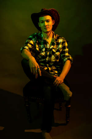 A young man in a plaid shirt with a cowboy hat sits on a chair, illuminated by a green and yellow lightの写真素材