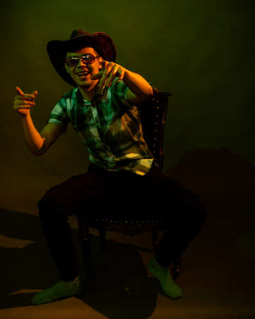 A young man in a plaid shirt with a cowboy hat sits on a chair, illuminated by a green and yellow...