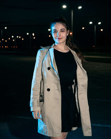 Girl in beige coat and dress against the backdrop of the night cityの写真素材