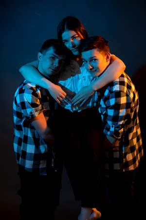 Two guys in a plaid shirt are holding a girl on a dark background in a studio with multicolored lightの写真素材