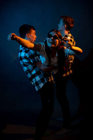 Two guys in a plaid shirt are holding a girl on a dark background in a studio with multicolored light.の写真素材