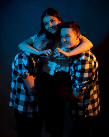 Two guys in a plaid shirt are holding a girl on a dark background in a studio with multicolored lightの写真素材