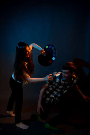A girl paints a guy in the studio on a dark background illuminated by multicolored lightの写真素材