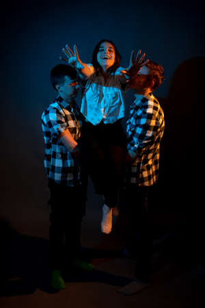 Two guys in a plaid shirt are holding a girl on a dark background in a studio with multicolored lightの写真素材