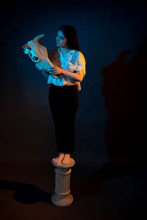 A girl stands on a Greek column with a bull skull in her hands illuminated by blue and yellow lightの写真素材