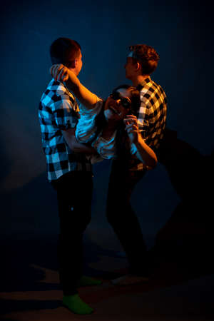 Two guys in a plaid shirt are holding a girl on a dark background in a studio with multicolored light.の写真素材