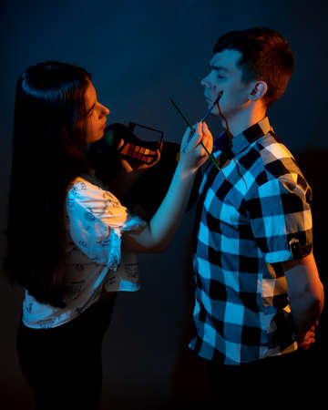 A girl paints a guy in the studio on a dark background illuminated by multicolored lightの写真素材