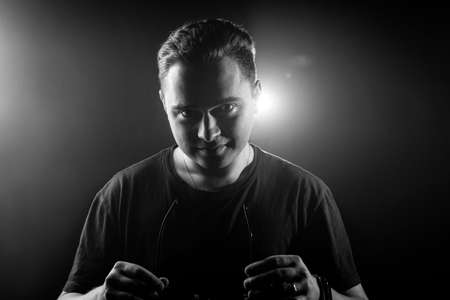 Black-and-white portrait of a young man holding glasses on a dark background with a glare behind his...