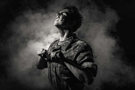 Black white portrait of a young soldier in combat coloring, scars and bruises, holding a machete on a black background with smokeの写真素材