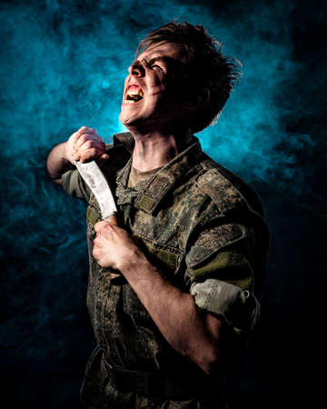 A large portrait of a soldier in scars and combat paint, holding a machete and screaming against the backdrop of blue smokeの写真素材