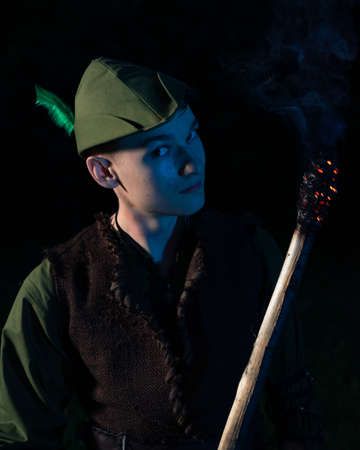 A guy dressed as Peter Pan stands with a burning torch against the night sky and dark forestの写真素材