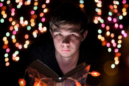 A young guy sits with a book in his hands, illuminated by colorful garlands in a fabulous atmosphere.の写真素材