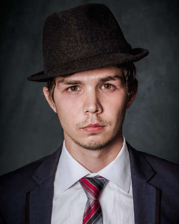 Portrait of a young guy in a black hat, white shirt, black jacket with red tie close-up on a dark background in the studioの写真素材