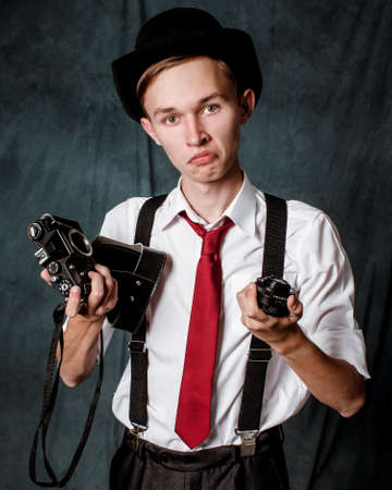 A young man in a white shirt, black hat and red tie with suspenders holds a disassembled camera in his hands and looks into the camera with surprise.の写真素材