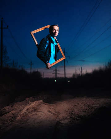 A guy peeks out of a frame hanging in the air at night on an empty dirt road.の写真素材