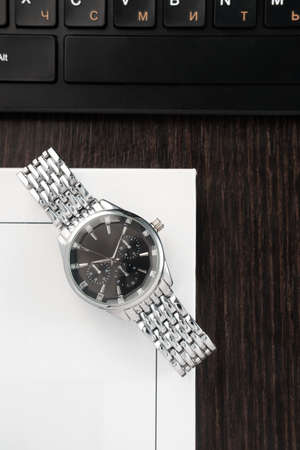 Wristwatch with a silver bracelet and a black dial lies on a book next to the keyboard on a dark wood tableの写真素材