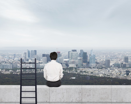 businessman in suit sitting on wall and looking to cityの写真素材
