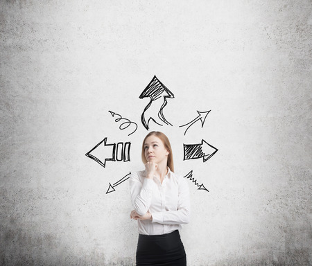 Beautiful blonde business woman is standing surrounded by arrows in different direction. A concept of decision making process. Concrete wall background.の写真素材