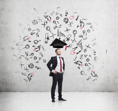 Young handsome businessman is thinking about education at business school. Drawn business icons over the concrete wall. Graduation hat.の写真素材