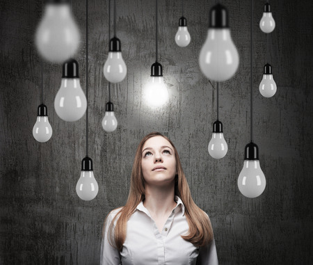 Charming lady is looking upward at the hanging light bulbs. A concept of searching new ideas. Dark concrete background.の写真素材