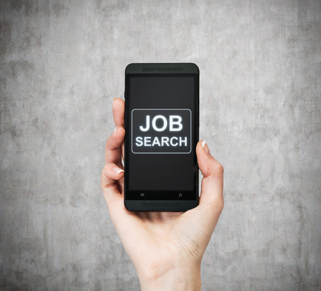 A hand holds a fancy smartphone. Words: ' Job search ' on the screen. A concept of recruitment. Concrete background.の写真素材