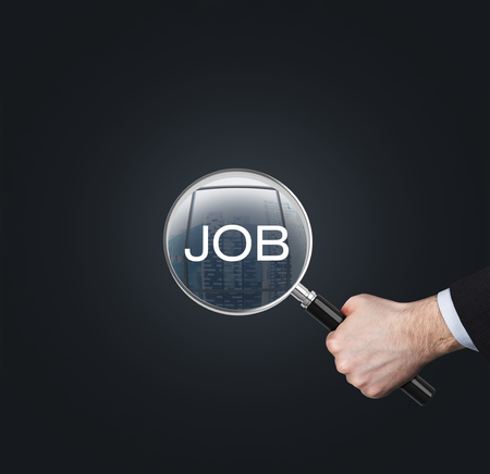 A hand holds a magnifying glass with the word 'JOB'. The concept of the searching of the job. Dark background.の写真素材